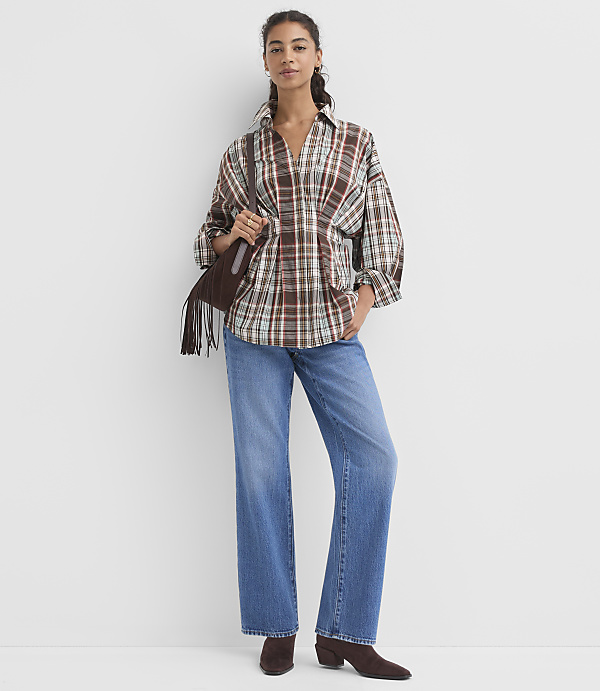 A woman in a brown plaid shirt and blue jeans stands against a plain white background, holding a fringed brown bag.