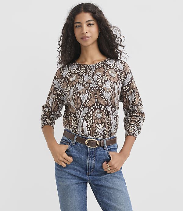 A woman in a patterned brown shirt and blue jeans stands against a plain gray background.