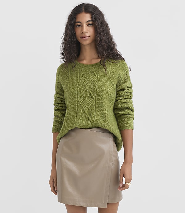 A woman in a green cable-knit sweater and a tan faux leather skirt stands against a plain gray background.
