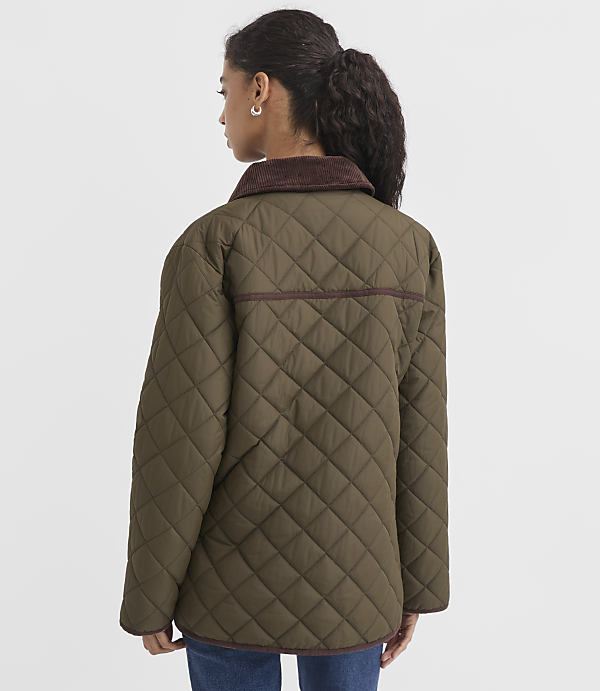 A woman in a quilted olive green jacket with a brown corduroy collar stands against a plain white background.