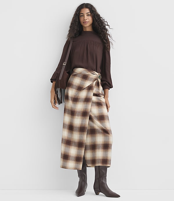 A woman in a brown smocked-neck blouse and a plaid wrap skirt stands against a plain white background.