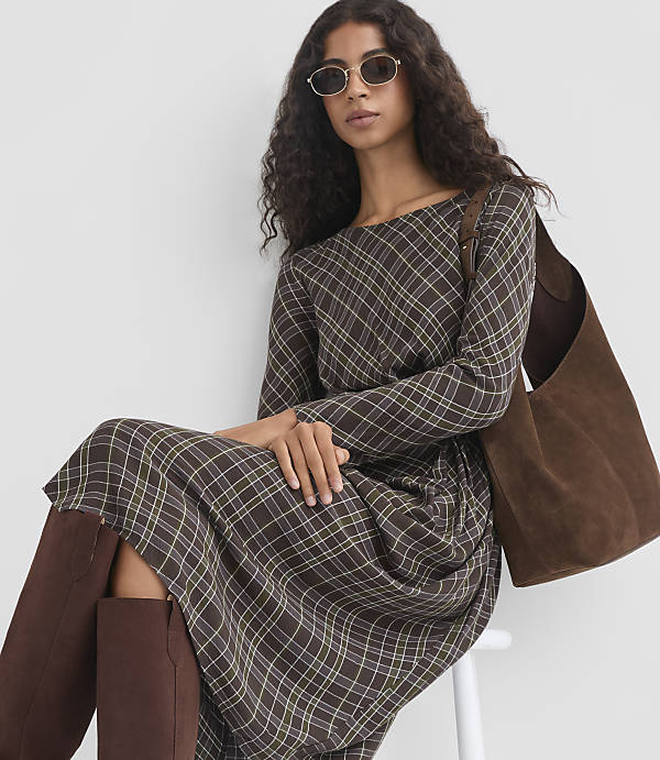 A woman in a plaid dress and brown boots sits in front of a white background, holding a brown suede bag.