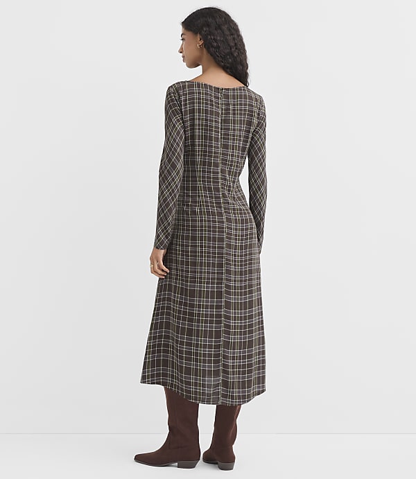 A woman in a long-sleeved plaid dress and brown boots stands against a plain white background.