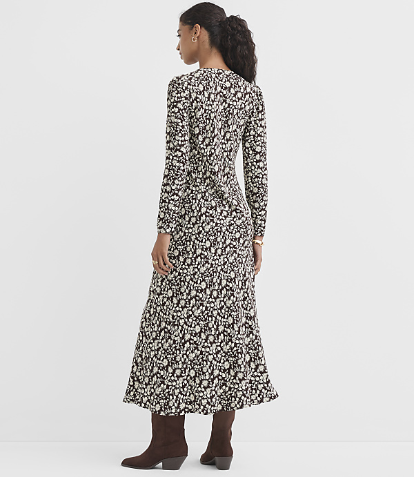A woman in a long-sleeved, floral-print dress and brown boots stands against a plain, light gray background.