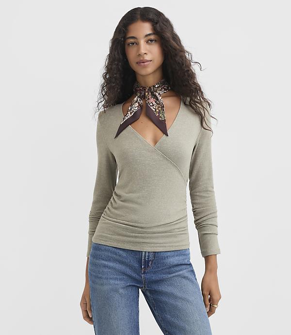 A woman in a light green wrap top and blue jeans stands against a plain gray background, a floral scarf tied around her neck.