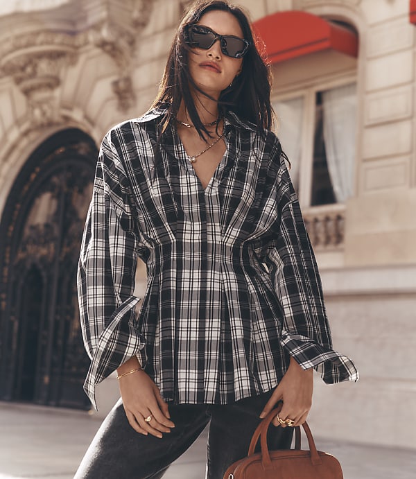 A woman in a plaid shirt and sunglasses stands in front of an ornate building.