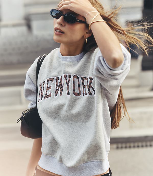 A woman in a grey sweatshirt with "NEW YORK" in leopard print stands in front of a blurred urban background.