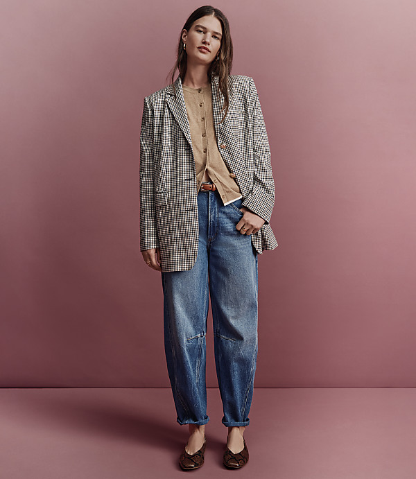 A woman in a checkered blazer, tan cardigan, and baggy jeans stands against a pink background.