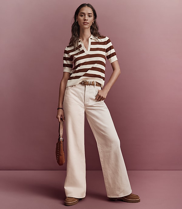 A woman in a striped polo shirt and wide-leg pants stands against a muted pink background.