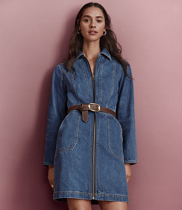 A woman in a denim dress with a brown belt stands against a pink background.