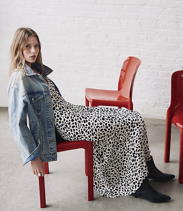 A young woman in a dalmatian print dress and denim jacket sits on a red chair in front of a white brick wall.
