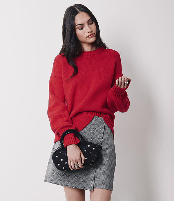 A woman in a red sweater and plaid skirt holds a studded black handbag against a plain white background.