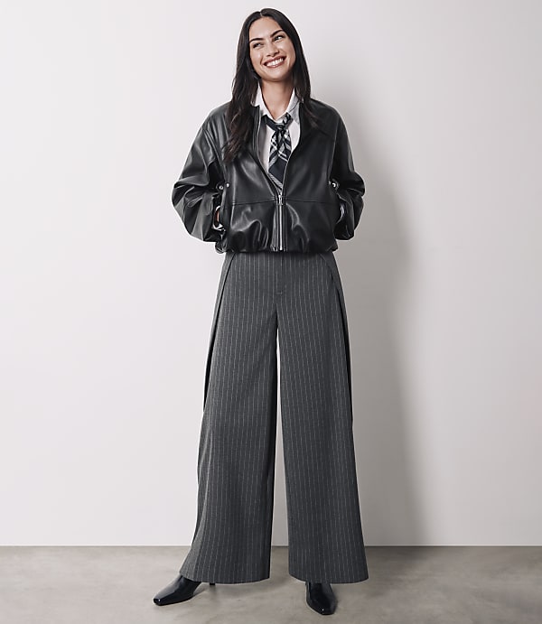 A smiling woman in a black leather jacket and pinstripe pants stands against a plain white background.