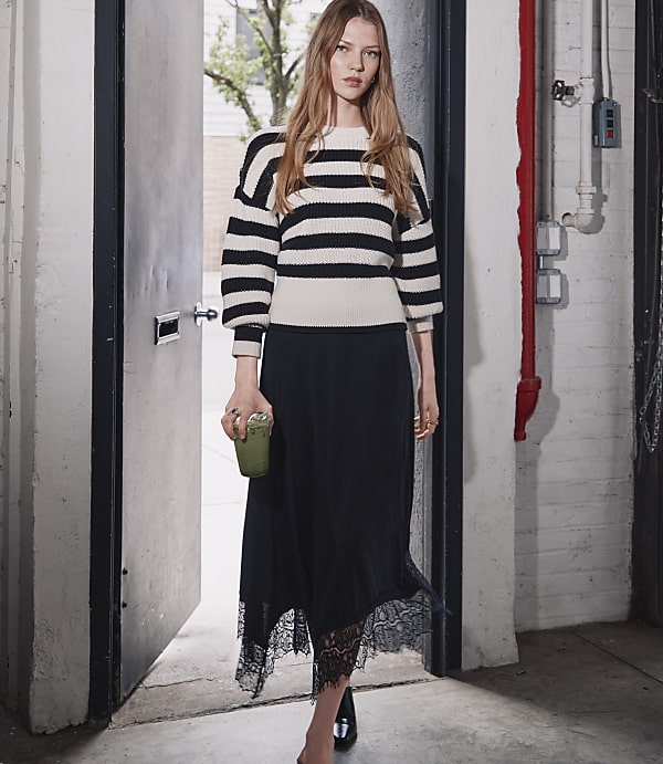 A woman in a striped sweater and black lace-trimmed skirt walks through a doorway, holding a green drink, with a brick building visible in the background.