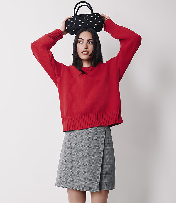 A woman in a red sweater and plaid skirt holds a patterned bag above her head against a plain white background.