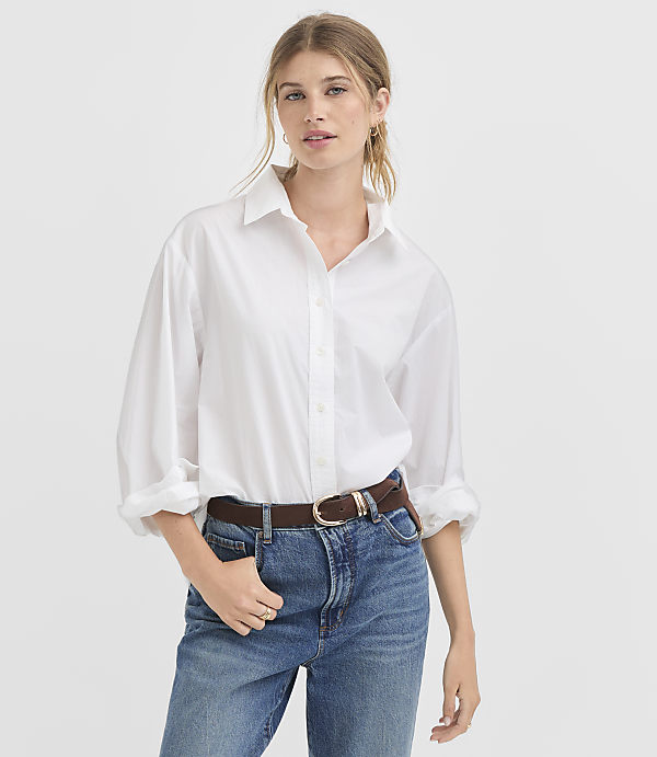 A woman in a white button-down shirt and blue jeans stands against a plain white background.