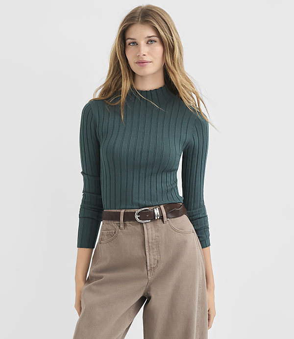 A woman in a teal ribbed sweater and brown pants stands against a plain white background.
