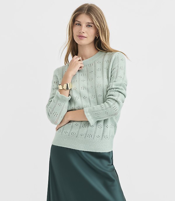A woman in a light green, textured sweater and a dark green satin skirt stands against a plain, light gray background.