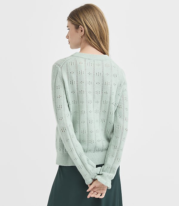 A woman in a light green, open-knit sweater stands with her back to the viewer against a plain white background.