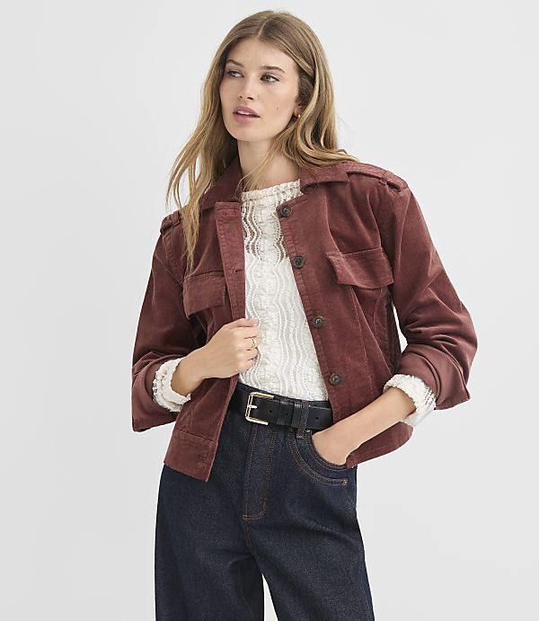 A woman in a rust-colored corduroy jacket and dark jeans stands against a plain white background.
