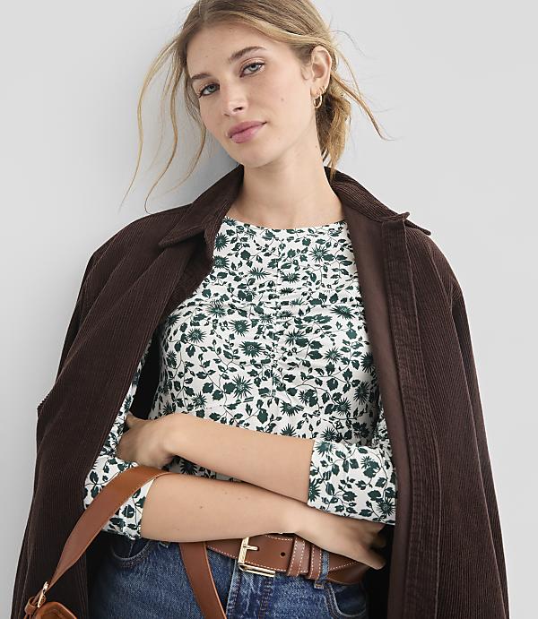 A woman in a floral top and brown jacket stands against a plain background.