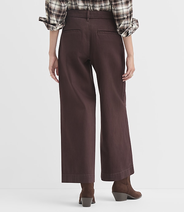A person wearing a plaid shirt and brown wide-leg pants stands against a plain white background.