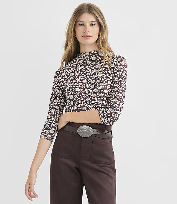 A woman in a floral mock-neck top and brown pants stands against a plain white background.