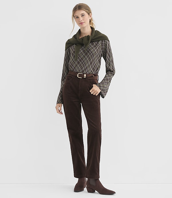 A woman in a brown plaid shirt, brown corduroy pants, and a green sweater draped over her shoulders stands against a plain white background.