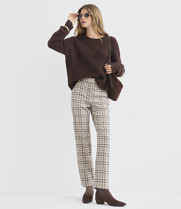 A woman in a brown cable-knit sweater and plaid pants poses against a plain white background.