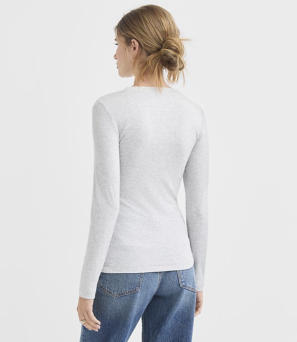 A woman in a grey long-sleeved shirt and blue jeans stands against a plain white background.