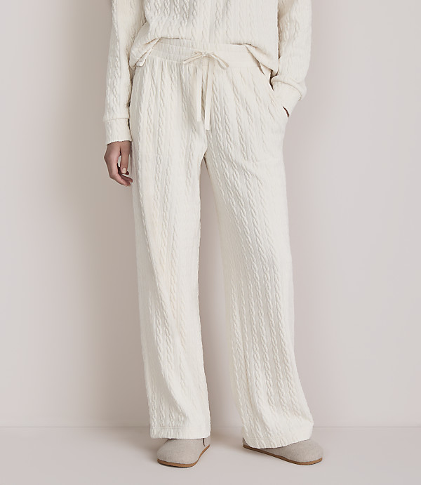 A person stands in cream-colored cable-knit pajamas and slippers against a plain, light-colored background.