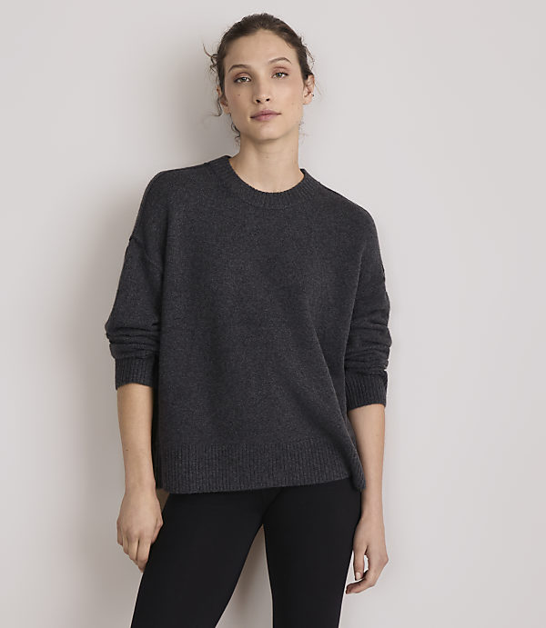 A woman in a dark grey sweater and black pants stands against a plain, light grey background.
