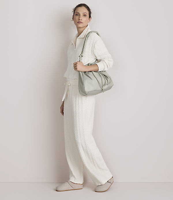 A woman in a cream-colored loungewear set and slippers walks with a light green drawstring bag against a plain, light-colored background.