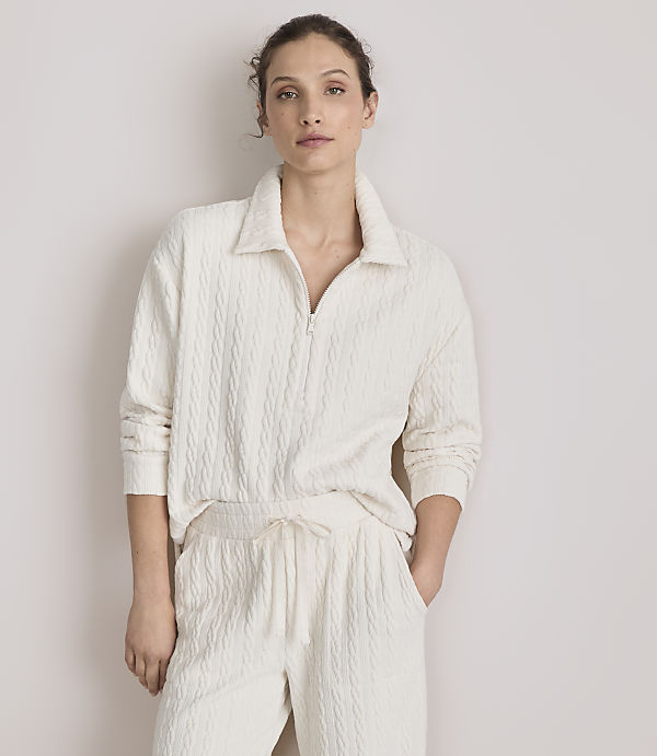 A woman in a cream-colored cable-knit loungewear set stands against a plain, light-colored wall.