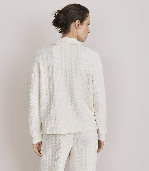 A woman in a cream cable-knit sweater and pants stands against a plain, light-colored background.