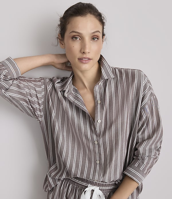 A woman in a striped pajama top rests her hand behind her head against a plain gray background.