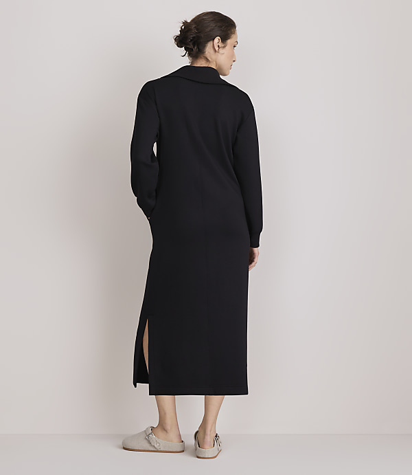 A woman in a black midi dress with side slits stands with her back to the camera, wearing light-colored slippers, against a plain, light-colored background.