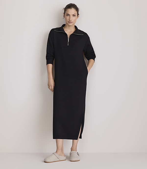 A woman in a black zip-neck dress and slippers stands against a plain, light-colored wall.