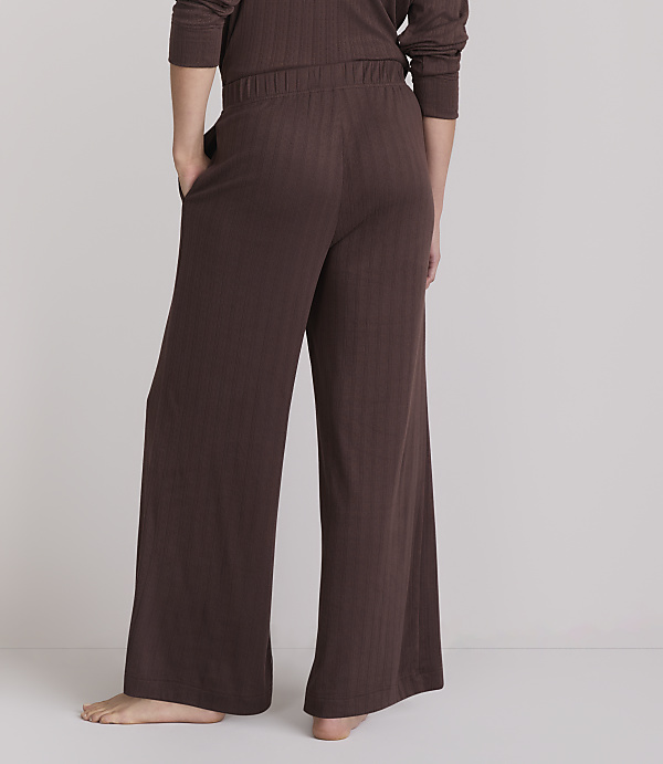 A person wearing brown ribbed pajama pants stands with their back to the camera against a plain, light gray background.