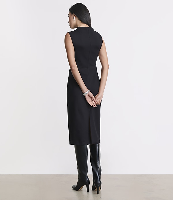 A woman in a black dress and boots stands with her hands behind her back against a plain, light gray background.