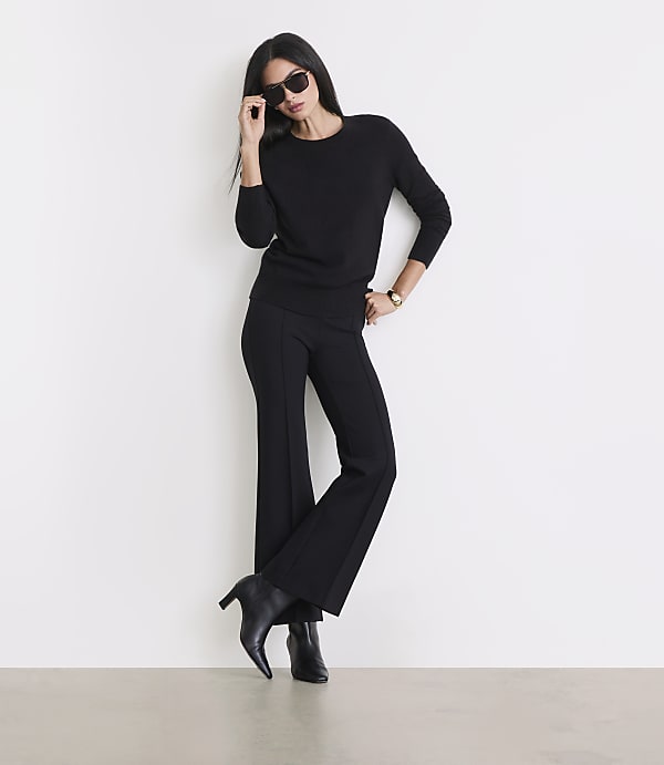 A woman in a black sweater and flared pants poses against a white wall, with a light-colored floor in the foreground.
