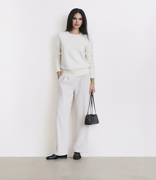 A woman in a cream sweater and matching pants stands against a plain white background, holding a small black purse.