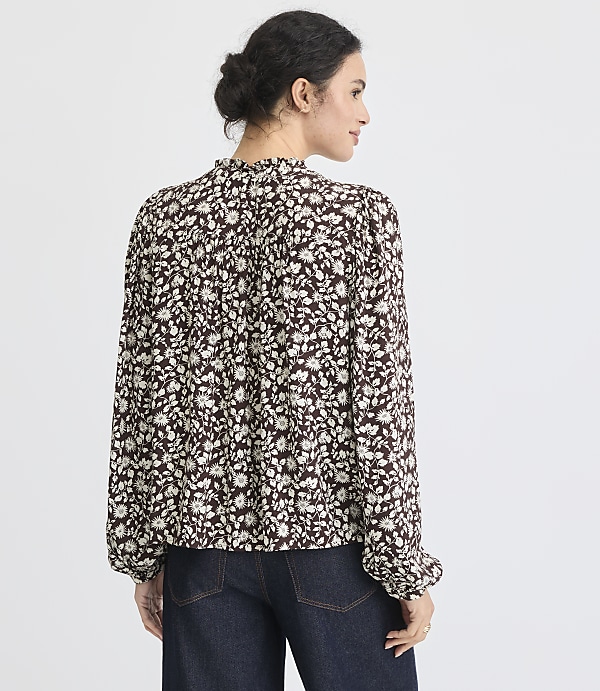 A woman in a floral blouse and dark jeans stands against a plain white background.