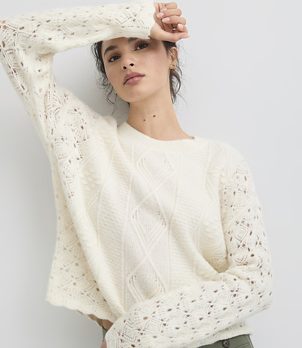 A woman in a cream-colored, open-knit sweater with her arm raised stands against a plain white background.