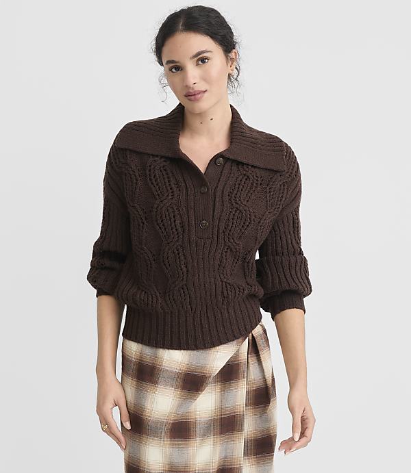 A woman in a brown cable-knit sweater and plaid skirt stands against a plain white background.