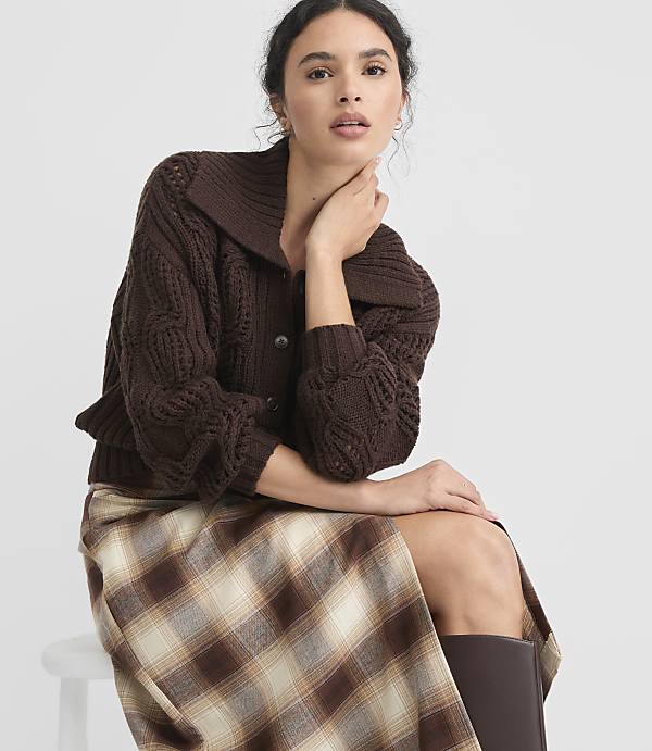 A woman in a brown cable-knit cardigan and plaid skirt sits against a plain white background.