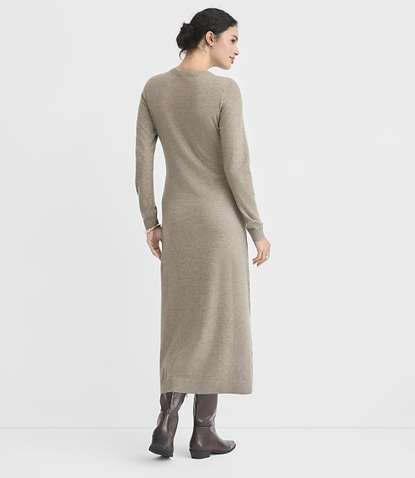 A woman in a long, taupe knit dress and brown boots stands against a plain, light gray background.