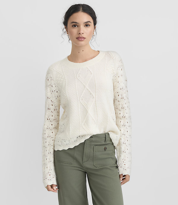 A woman in a cream cable-knit sweater and green pants stands against a plain white background.