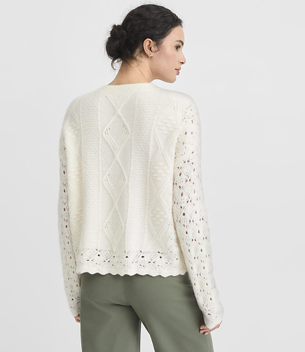 A woman in a cream-colored, cable-knit sweater and green pants stands against a plain gray background.