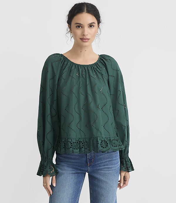 A woman in a green eyelet blouse and blue jeans stands against a plain white background.